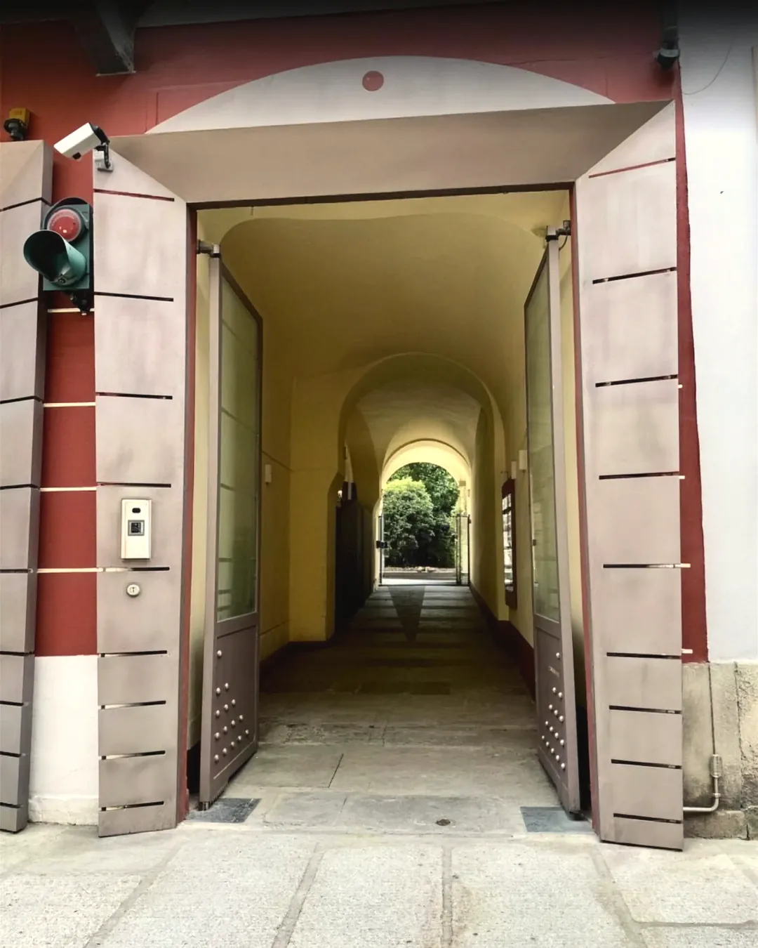 Il portone di via Donizetti 4 a Varese, aperto: i battenti grigi sull’intonaco rosso, l’androne ad archi e in fondo il verde del cortile.