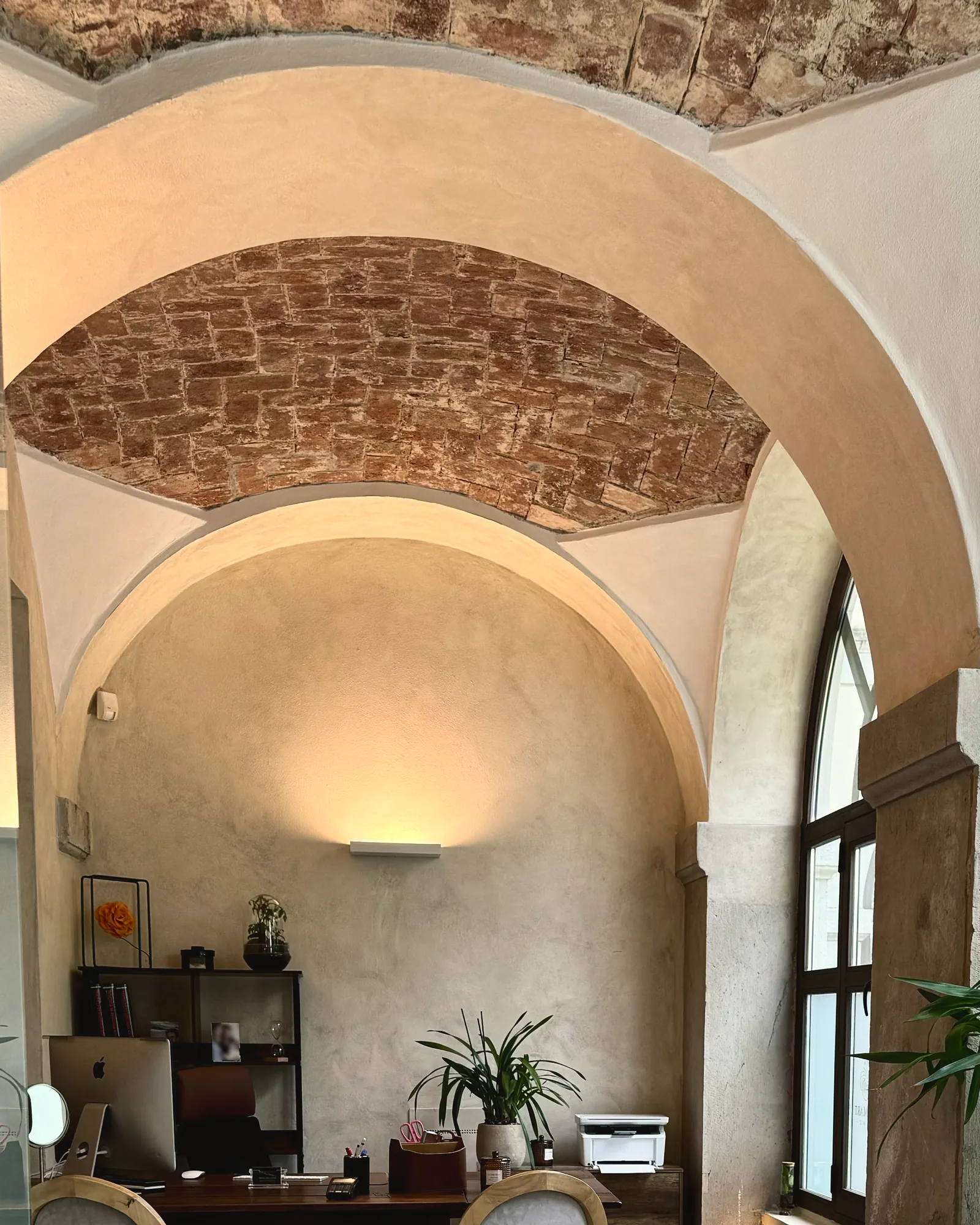 Lo studio di Derm Art Clinic in via Donizetti 4: la volta in mattoni, gli archi chiari e la luce calda di una lampada sulla parete, sopra la scrivania.